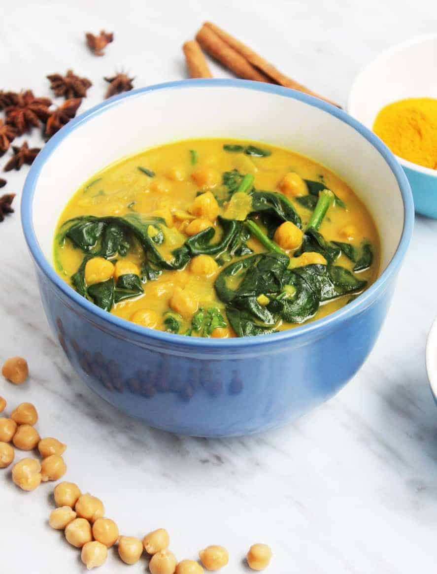 A big bowl of chickpea & spinach curry on a white marble tabletop, styled with the ingredients used in the dish