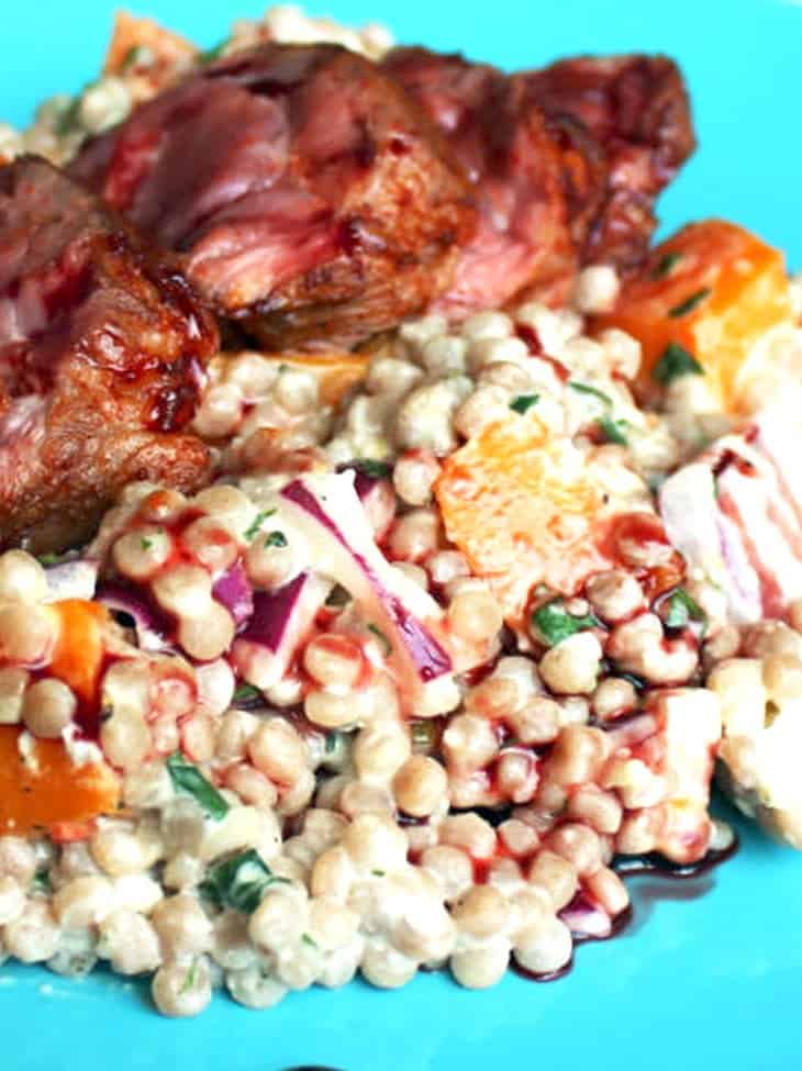 Giant couscous salad on a blue plate, topped with sliced lamb neck