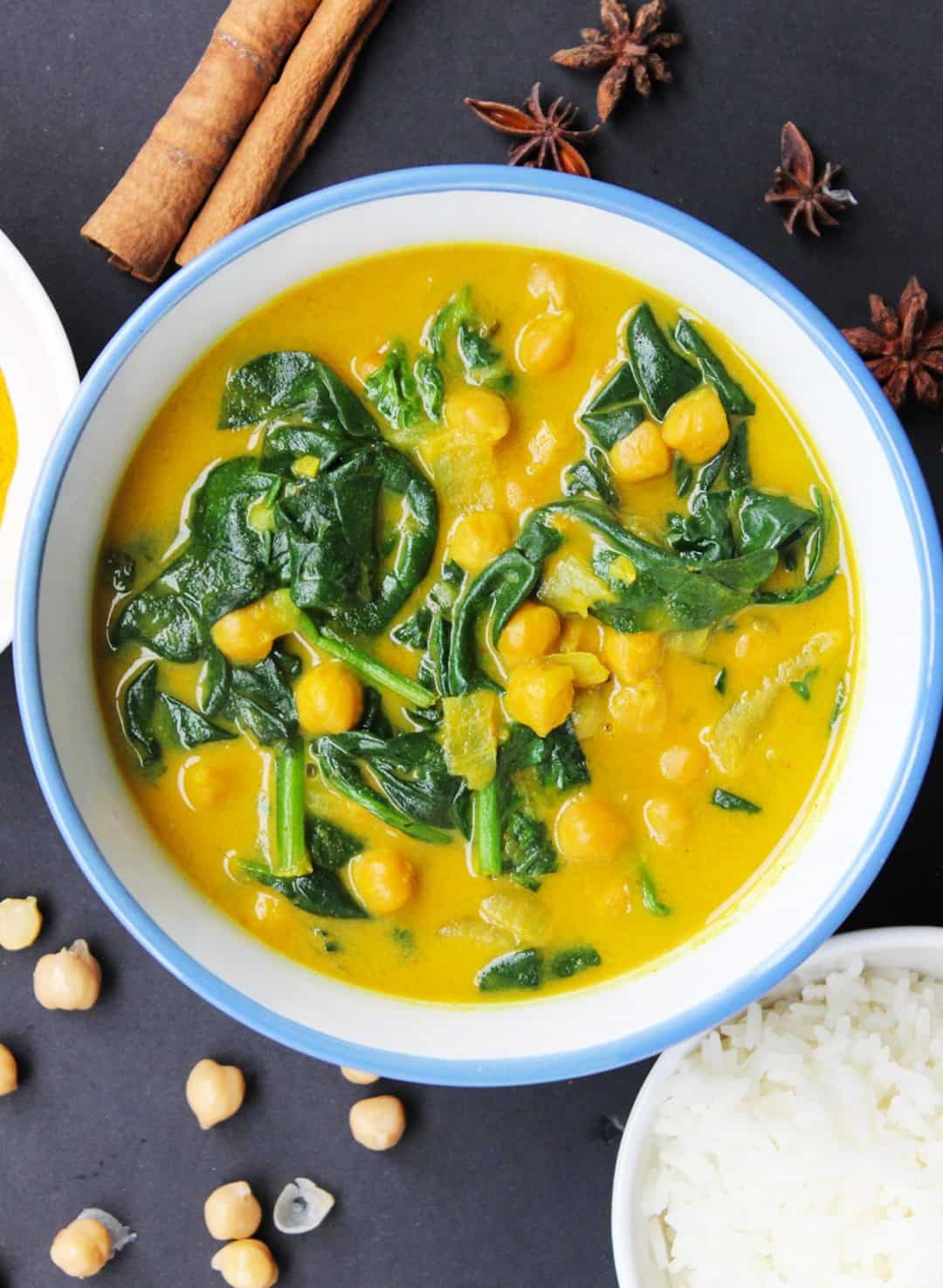 A bowl of chickpea & spinach curry on a black tabletop, styled with the ingredients used in the dish