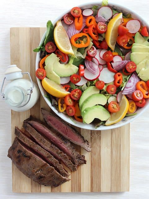 A bowl of salad and sliced flank steak and a bottle of dressing on a chopping board
