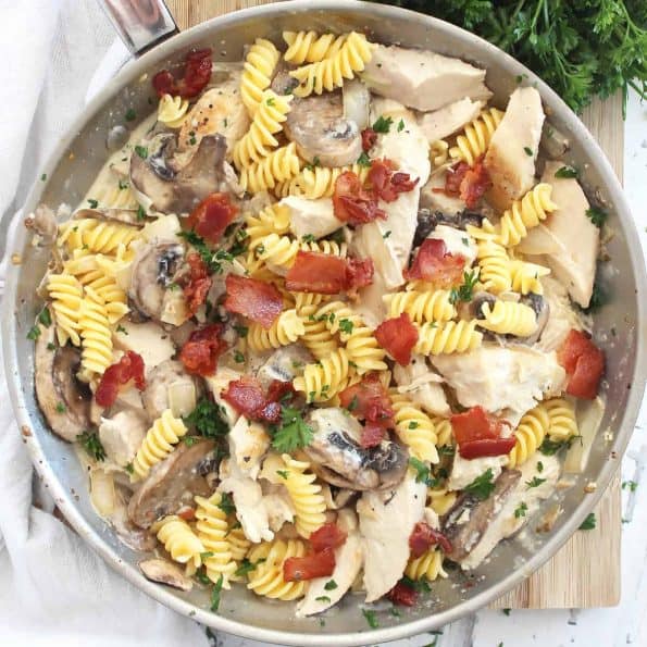 A silver skillet with chicken and mushroom pasta on a wooden chopping board.