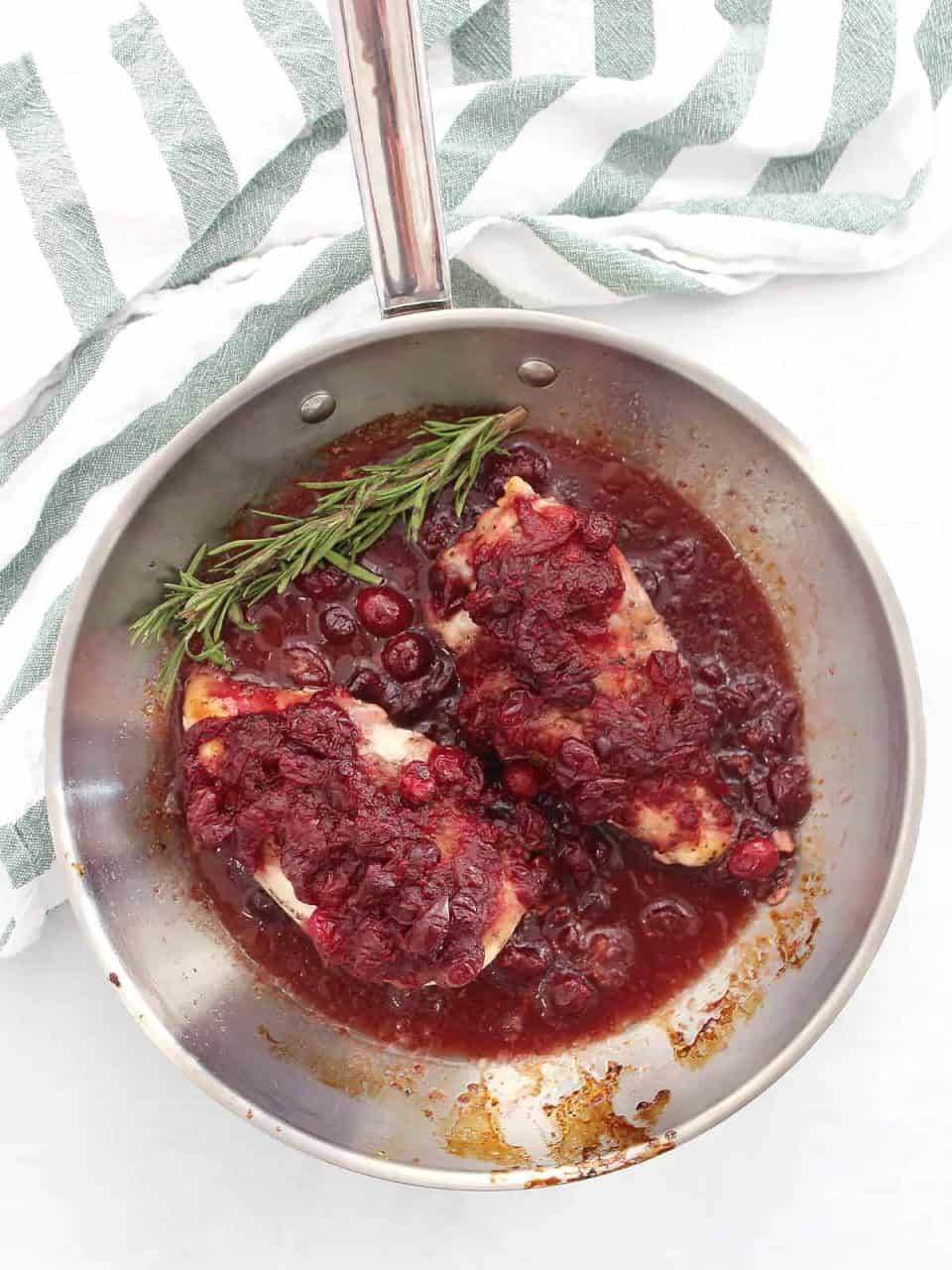 Two cranberry covered chicken breasts in a skillet with sauce and fresh rosemary.