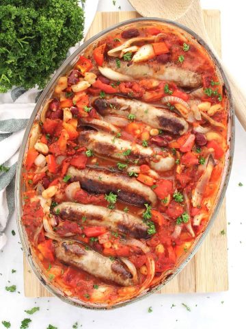 Sausage and bean casserole in a baking dish next to a wooden spoon and fresh herbs.