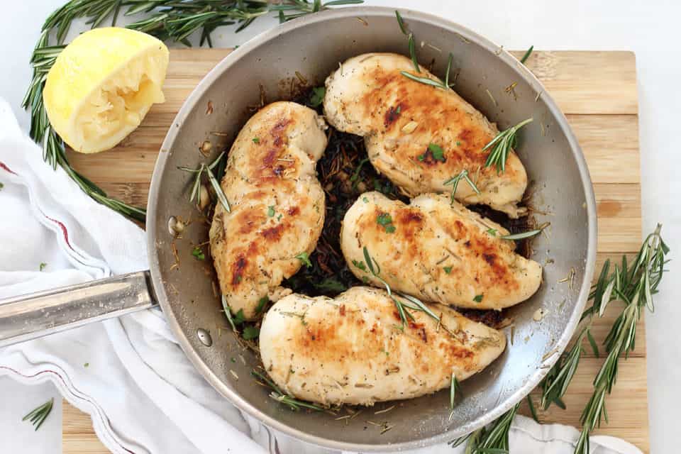 Four chicken breasts cooked in a skillet next to fresh lemon and rosemary.