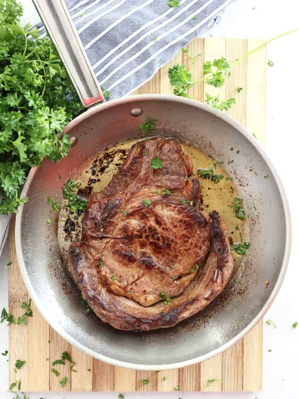 Beer marinated steak in a metal skillet on a wooden chopping board next to a bunch of fresh parsley.