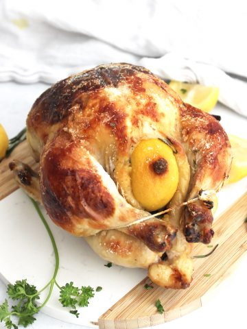A whole roasted buttermilk chicken with legs trussed together next to fresh herbs and lemons on a chopping board.