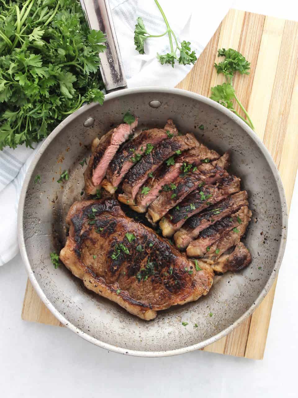 Two reverse seared cola marinated steaks in a skillet with one cut into slices and garnished with parsley.