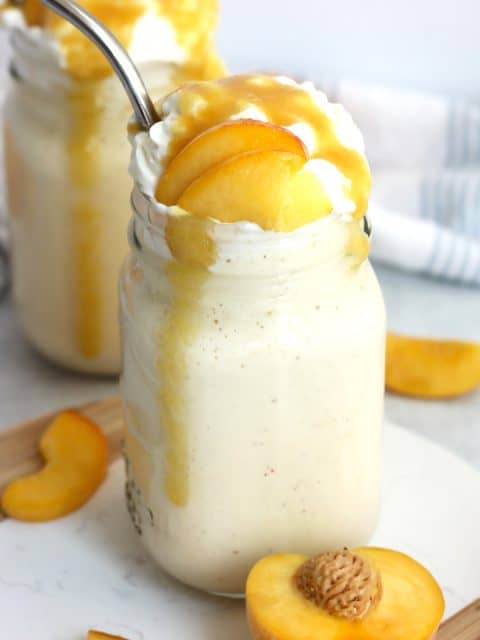 A peach milkshake in a glass jar with whipped cream, peach slices and a metal straw.