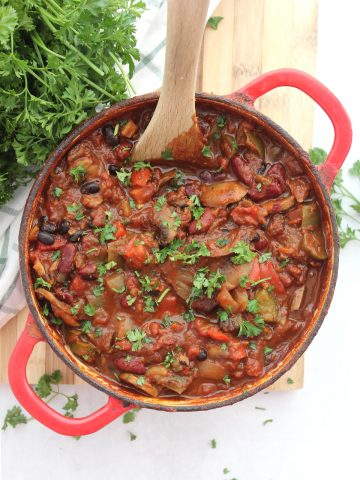 Vegan mushroom chili in a red Dutch oven with a wooden spoon and garnished with fresh herbs.