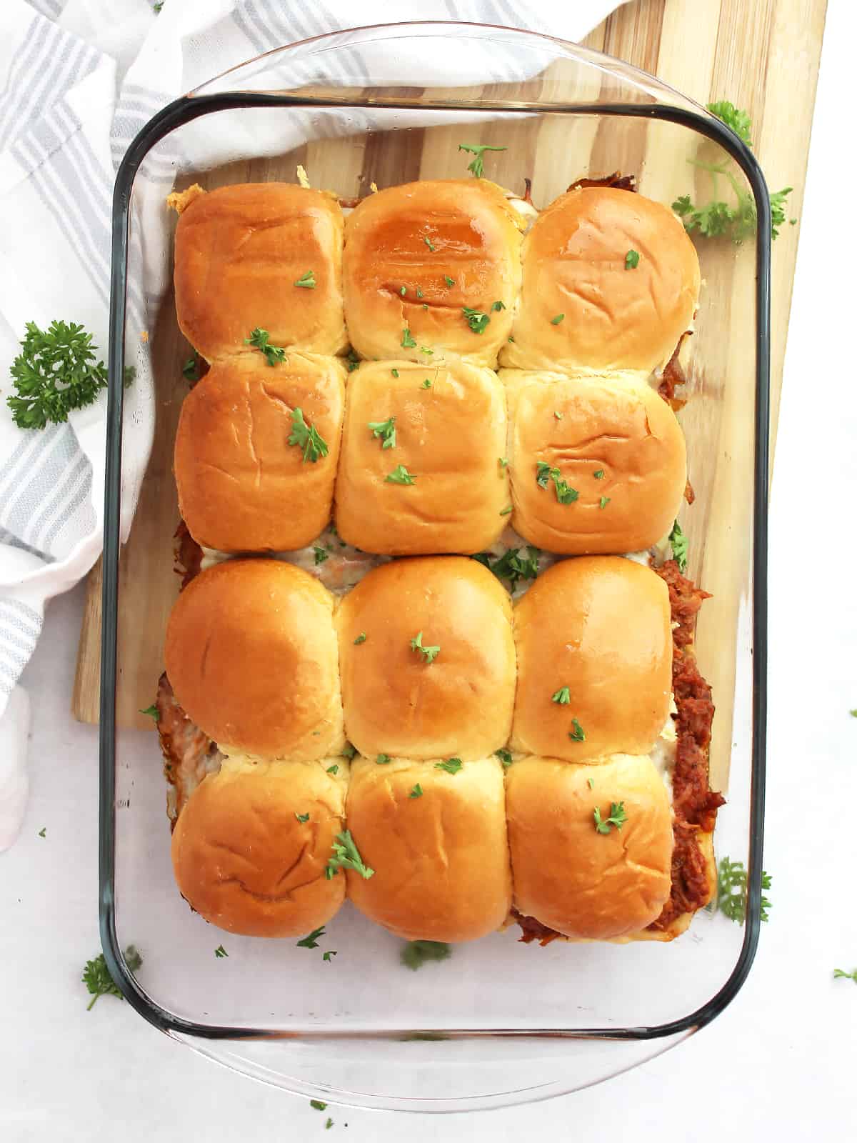Overhead shot of twelve pulled pork sliders in a glass casserole dish garnished with fresh parsley.