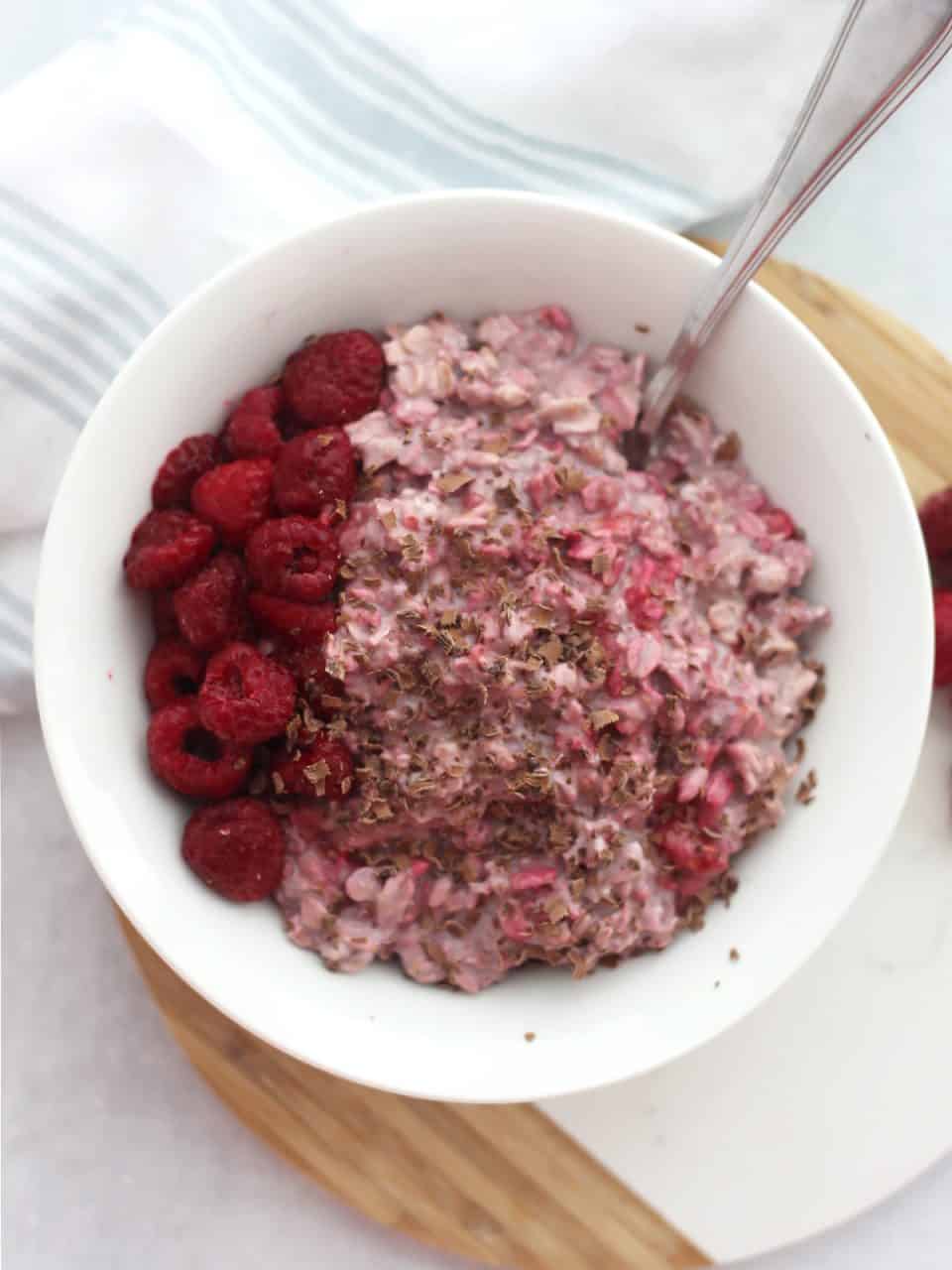 A spoon in a bowl of overnight oats with fresh raspberries and shaved dark chocolate.
