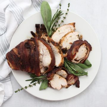 Carved bacon wrapped turkey breast on a serving plate garnished with fresh herbs.