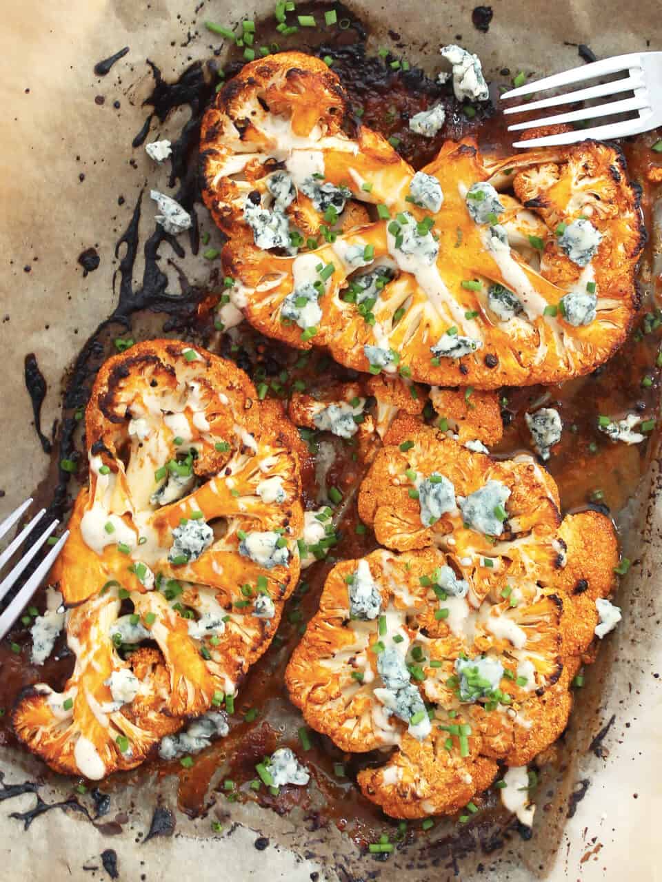 Overhead shot of three buffalo cauliflower steaks topped with blue cheese and chives on a parchment lined baking sheet with two forks.