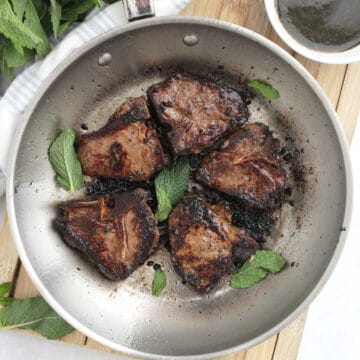 Five seared lamb chops in a silver skillet with fresh mint leaves.
