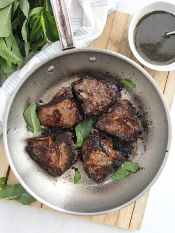 Five reverse seared lamb chops in a skillet with fresh mint leaves and next to a bowl of mint sauce.