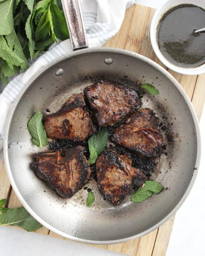 Five reverse seared lamb chops in a skillet with fresh mint leaves and next to a bowl of mint sauce.