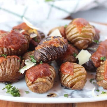 Mini roasted hasselback potatoes on a plate garnished with shredded parmesan and fresh herbs.
