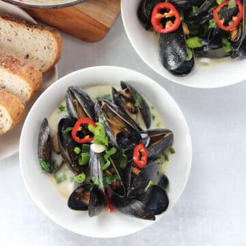 A bowl of cooked mussels in Thai coconut milk garnished with red chilies and green onion.