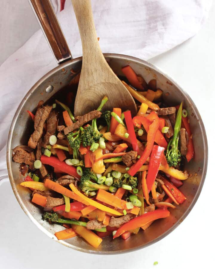 Stir fried beef strips with vegetables in a silver skillet with a wooden spoon.