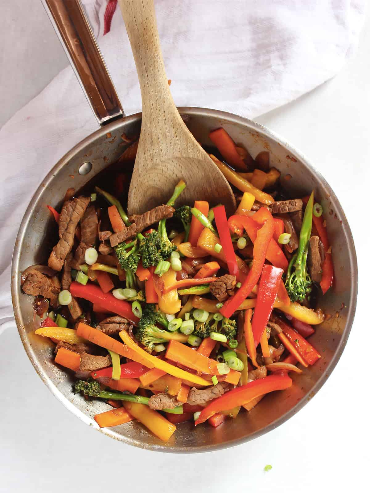 Stir fried beef strips with vegetables in a silver skillet with a wooden spoon.