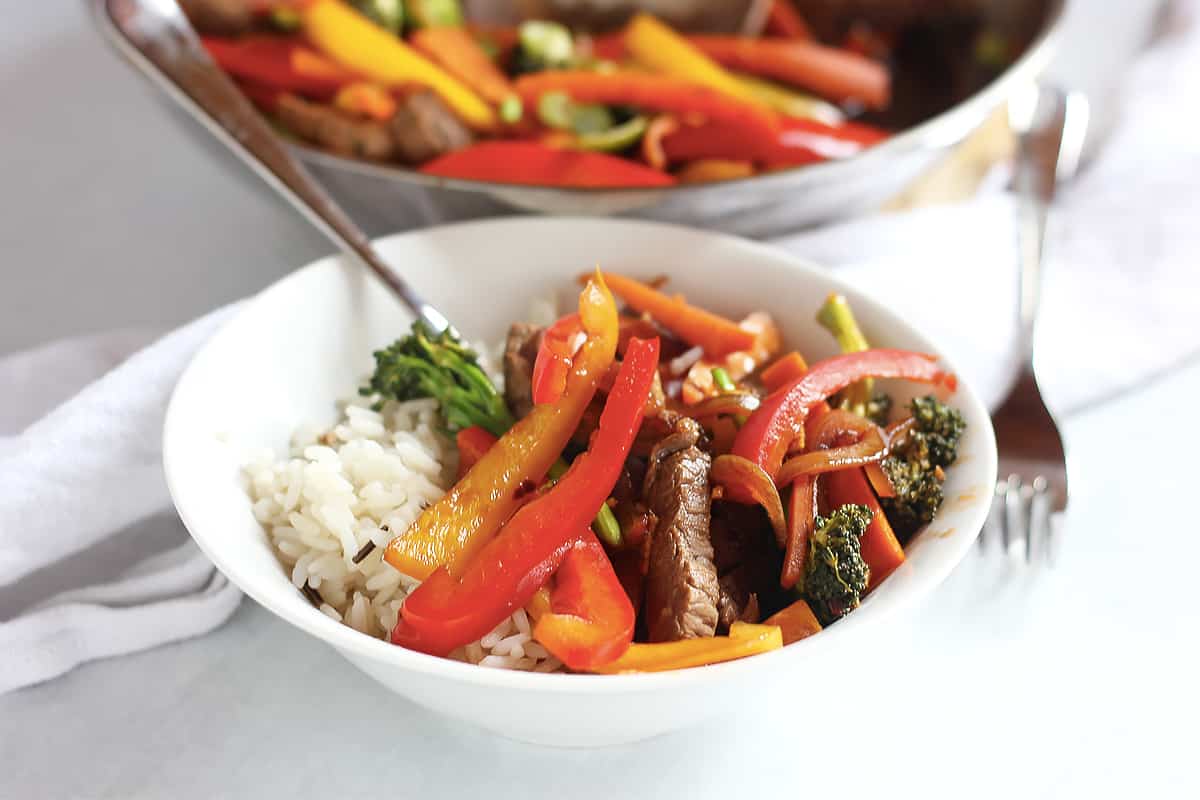 Marinated beef and vegetable stir fry served over rice in a white bowl.