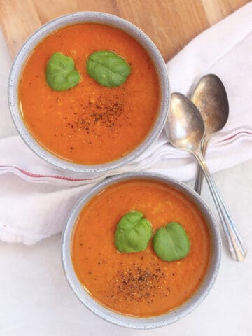 Two bowls of tomato and vegetable soup topped with fresh basil leaves and black pepper.