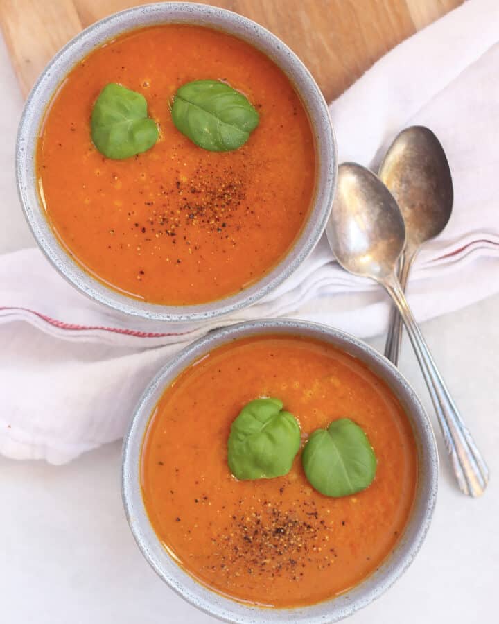 Two bowls of tomato and vegetable soup topped with fresh basil leaves and black pepper.