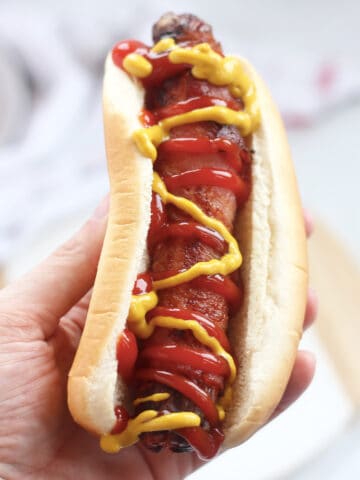 A hand holding a bacon wrapped hot dog.