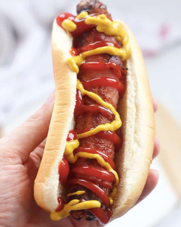 A hand holding a bacon wrapped hot dog.
