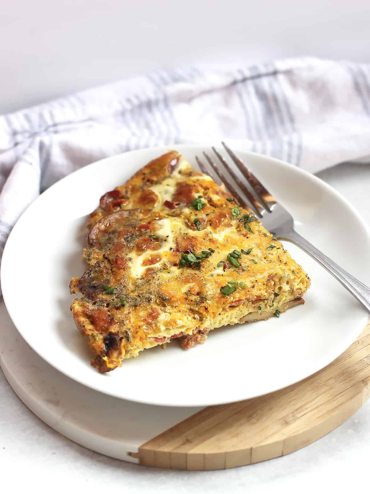 A slice of egg bacon frittata served on a white plate with a fork.