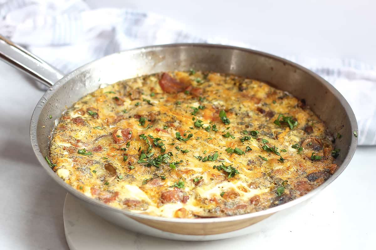 A bacon, mushroom and cheese frittata in a skillet on a white chopping board.