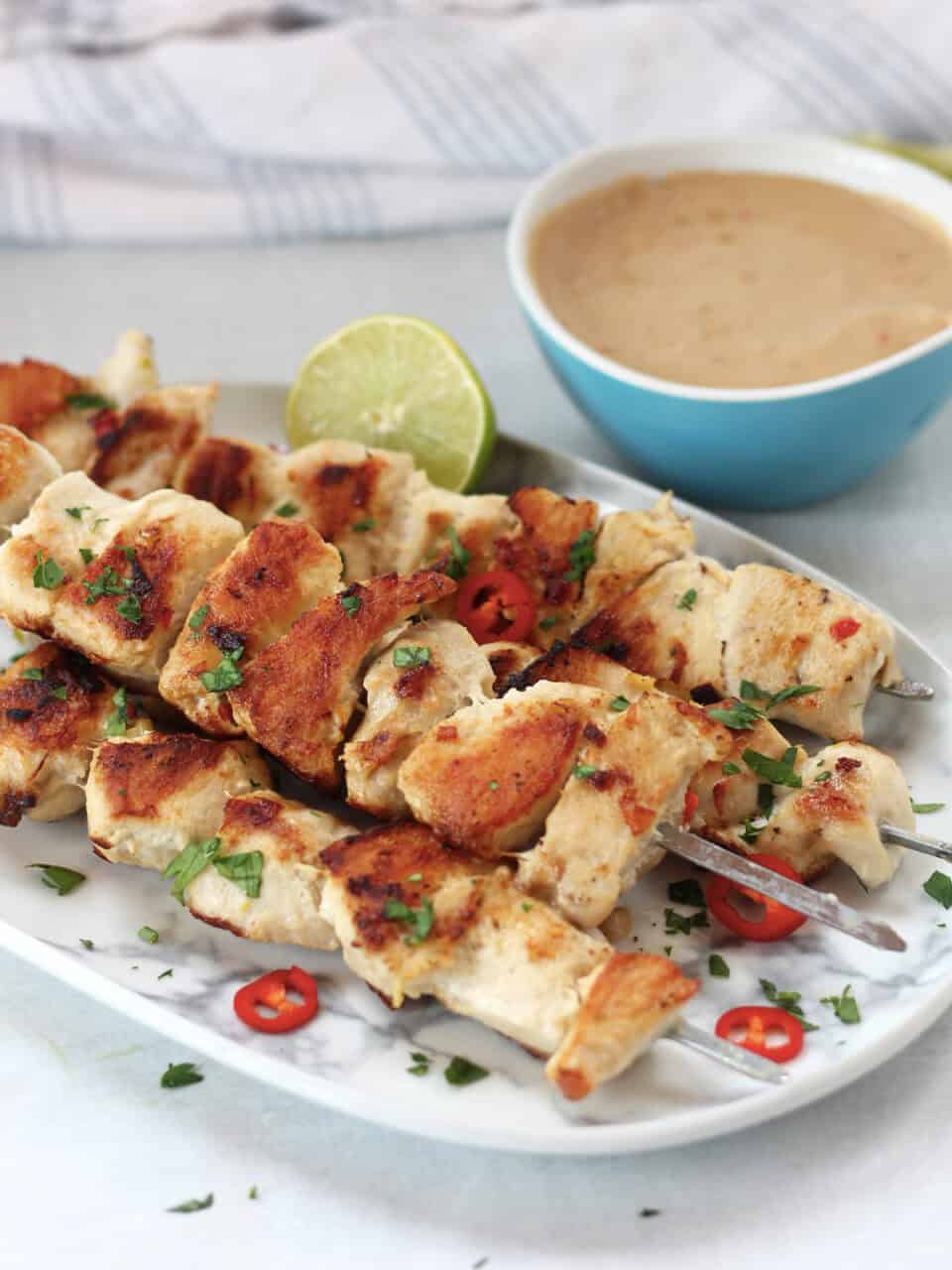 Browned chicken on metal skewers next to a bowl of peanut dip.