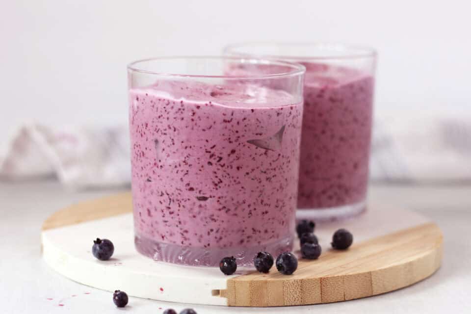 Two glasses of purple milkshake next to fresh blueberries.