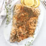 A baked fillet of salmon with thyme seasoning on a serving plate next to lemon slices.