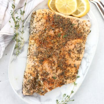A baked fillet of salmon with thyme seasoning on a serving plate next to lemon slices.