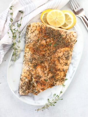 Cooked salmon fillet on a plate next to fresh thyme sprigs and lemon slices.