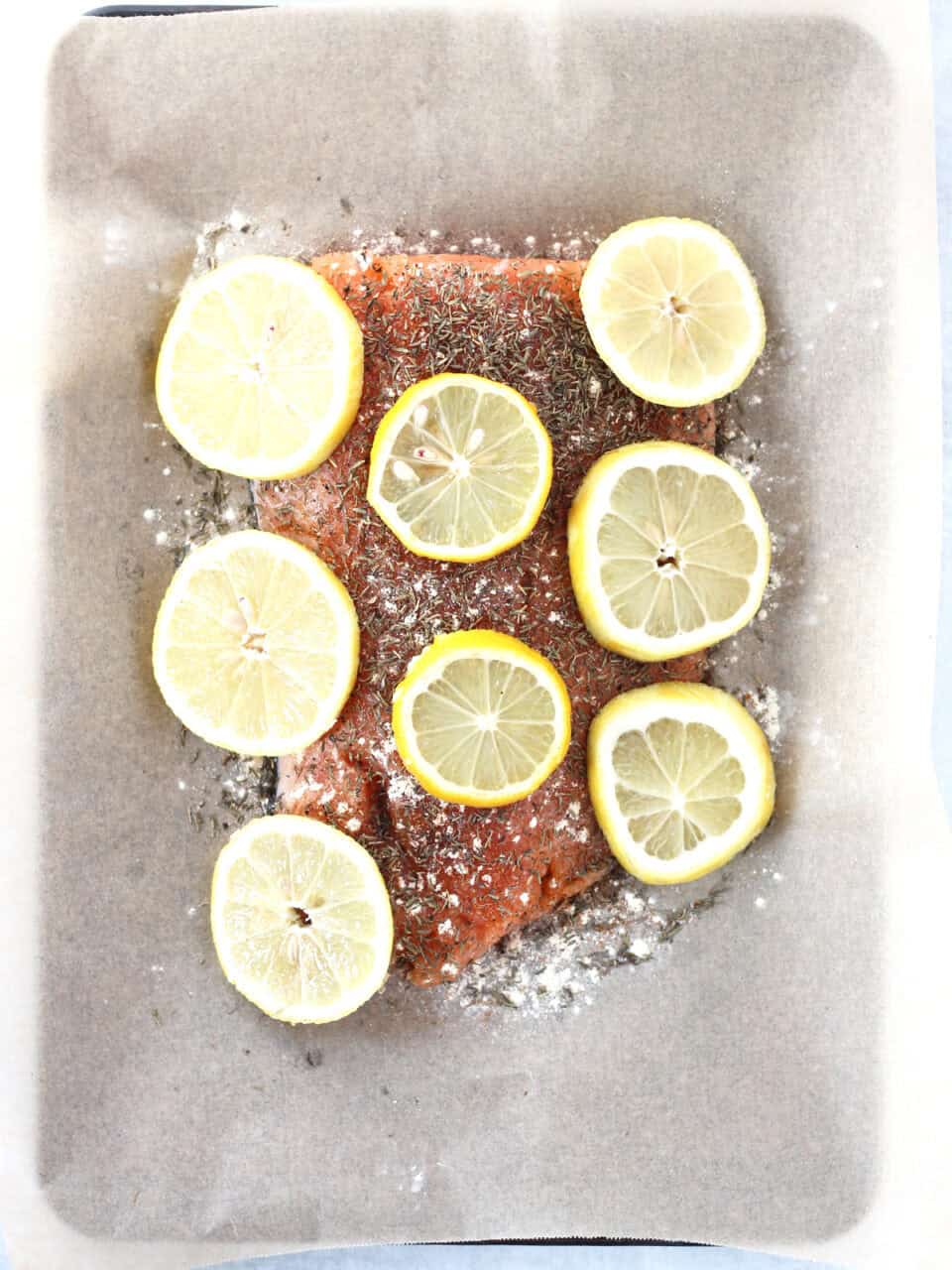 A raw salmon fillet on parchment with seasoning sprinkled on top and lemon slices placed on top.