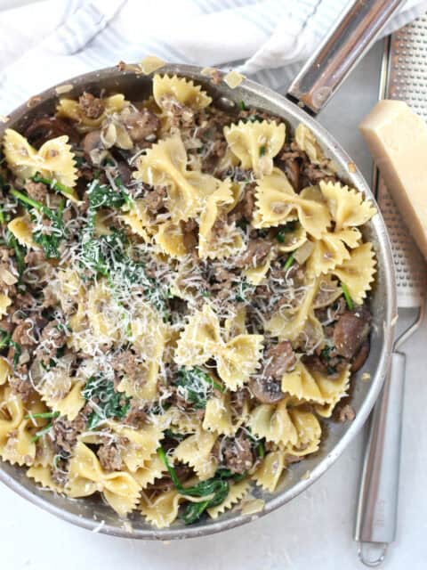 Bowtie pasta with mushrooms, beef and spinach, topped with parmesan, in a silver skillet.