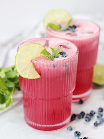A pink drink in two tall glasses next to fresh mint, blueberries and a halved lime.