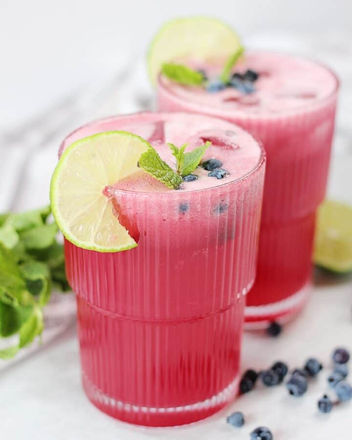 A pink drink in two tall glasses next to fresh mint, blueberries and a halved lime.