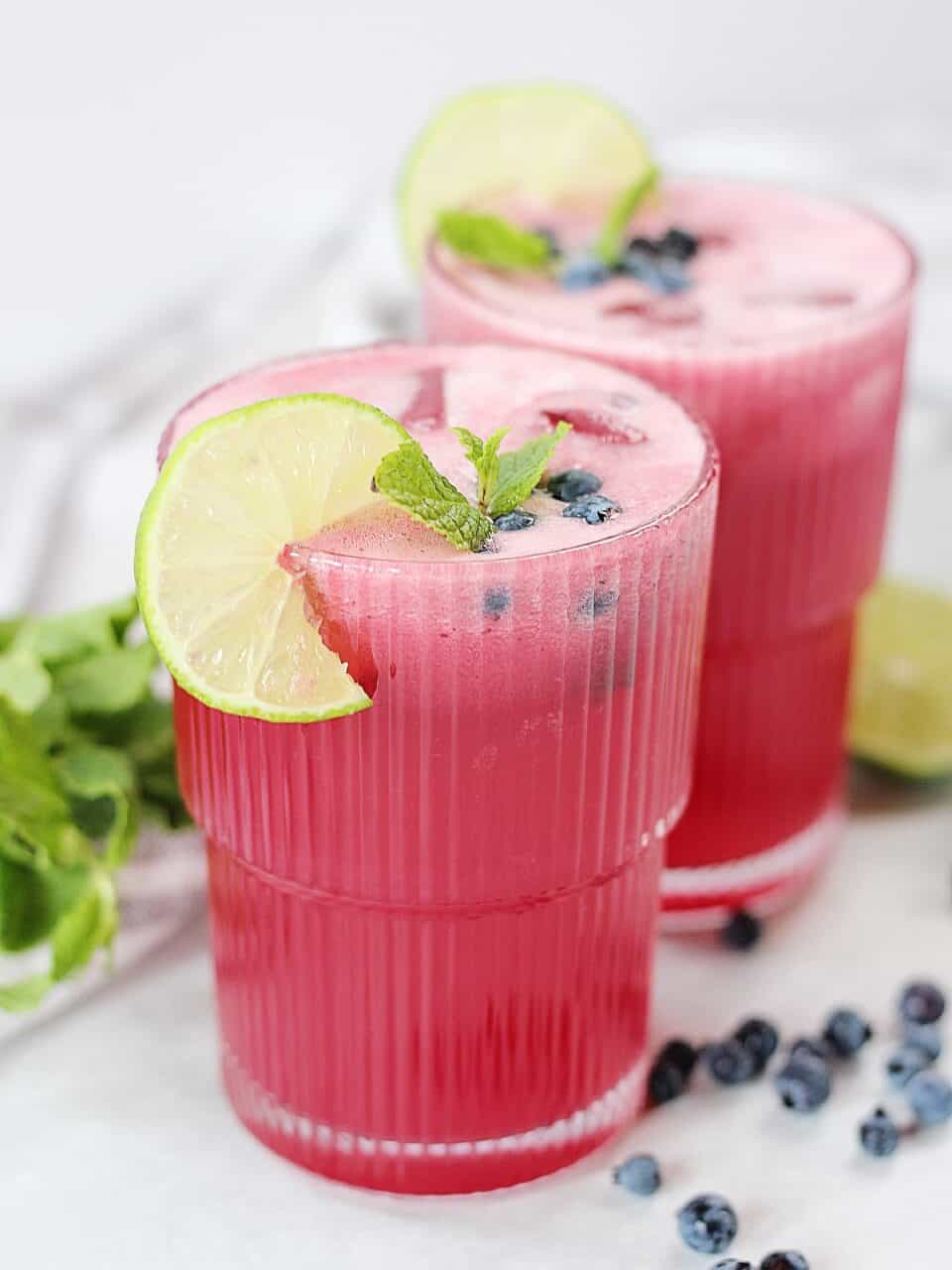 A pink drink in two tall glasses next to fresh mint, blueberries and a halved lime.